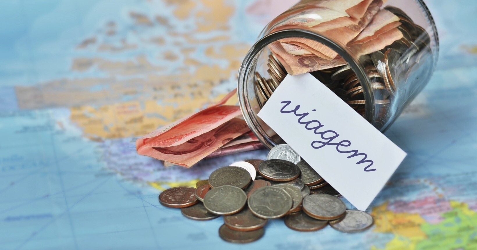 Cofrinho com moedas e cédulas sobre um mapa com etiqueta escrita 'viagem', simbolizando planejamento financeiro para viajar mesmo com o dólar e IOF mais caro